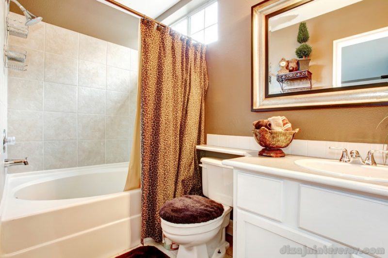 Beige wall bathroom with white washbasin cabinet, mirror, toilet and tub. Decorated with leopard print curtains, brown toilet cover and soft rug