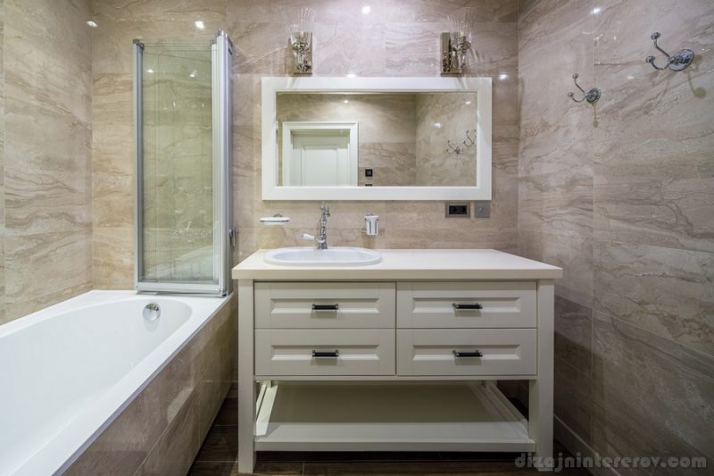 Stylish luminous bathroom in a classic style with light textured walls. There is a white bath with glass folding partition, sink, white wooden lockers, mirror with two lamps hanging over it.